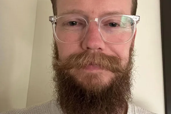 Jordon Leamaster headshot, inside wearing glasses and a grey shirt. Jordon has a long beard and mustache, combed back hair.
