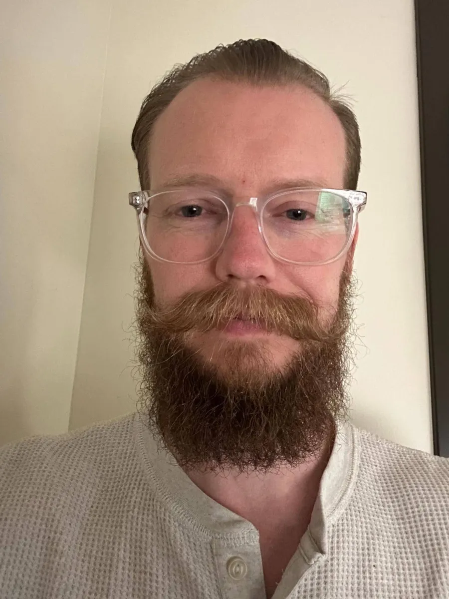 Jordon Leamaster headshot, inside wearing glasses and a grey shirt. Jordon has a long beard and mustache, combed back hair.